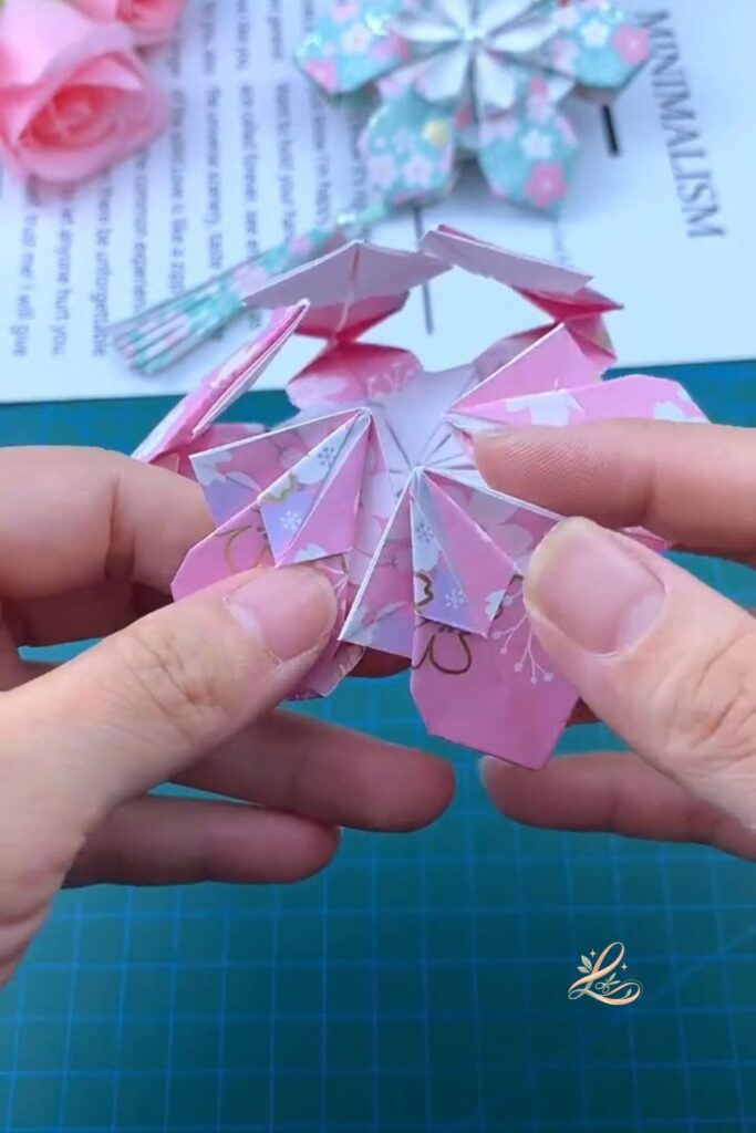 The partially assembled flower held in both hands, four or five modules joined and fanning outward in a clear arc, the flower shape emerging, the patterned paper petals clearly visible