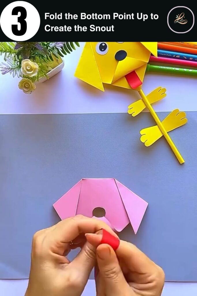 Pink paper folded into a dog face shape with ear flaps and snout, step in making a paper dog pencil topper